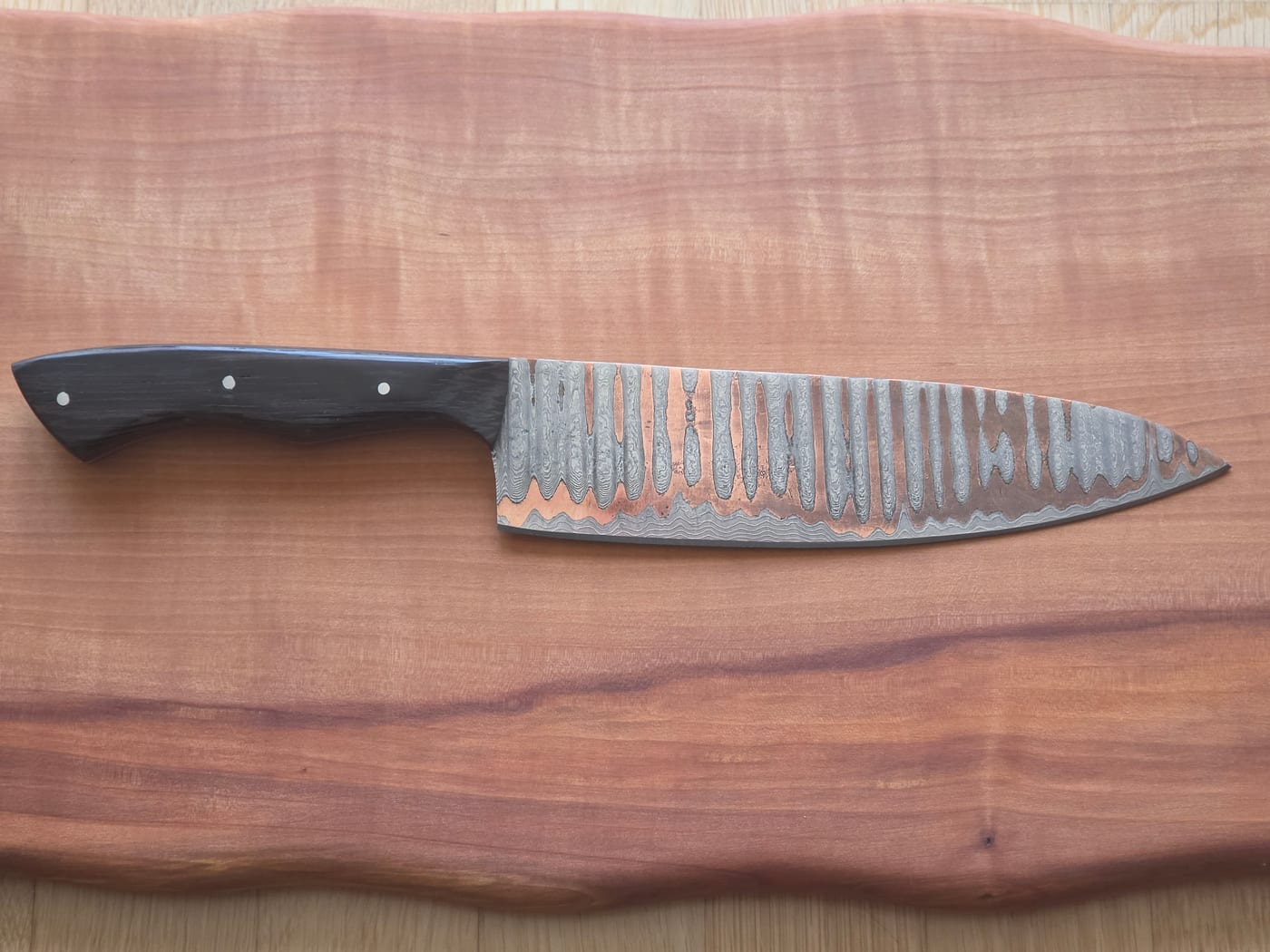 Kochmesser mit Damastklinge in Kupfertönen und dunklem Holzgriff auf einem Schneidbrett aus Kirschholz