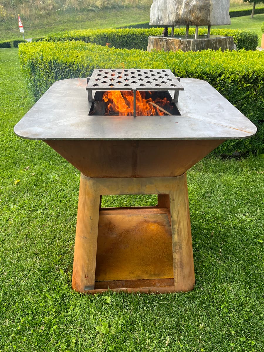 Feuerplatte aus Stahl mit offenem Feuer und abnehmbarem Rost, im Garten aufgestellt